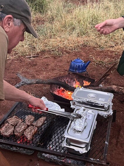 A person grilling steaks on an outdoor grill with foil-wrapped trays nearby. 4