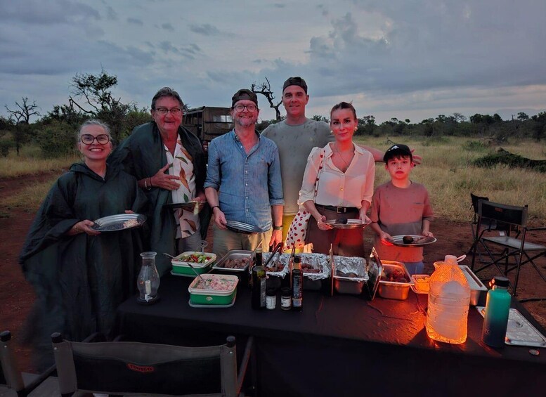 A group of people at an outdoor event with a table set up for a meal, featuring food containers, a water bottle, and a portable stove. 1