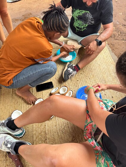 A group of people sitting on a woven mat outdoors, engaged in a food preparation activity with bowls and coconuts. 4