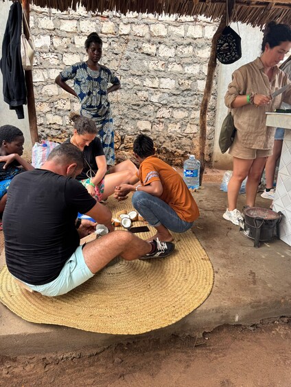 A group of people gathered around a table, some sitting and others standing, in an outdoor setting with a stone wall and thatched roof in the background. 2