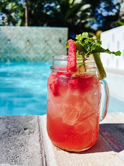 A mason jar with a red drink, mint garnish, and a straw, placed on a concrete surface by a pool. 2