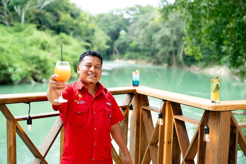 A person in a red shirt holding a glass of orange juice, standing on a wooden deck with a river and greenery in the background. 4
