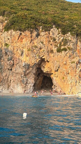 A rocky coastline with a cave entrance, a beach, and people swimming and sunbathing. 5