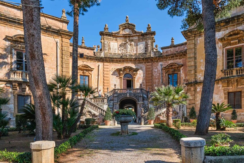 A historic building with ornate architecture, a central archway, and a well-maintained garden with palm trees. 1