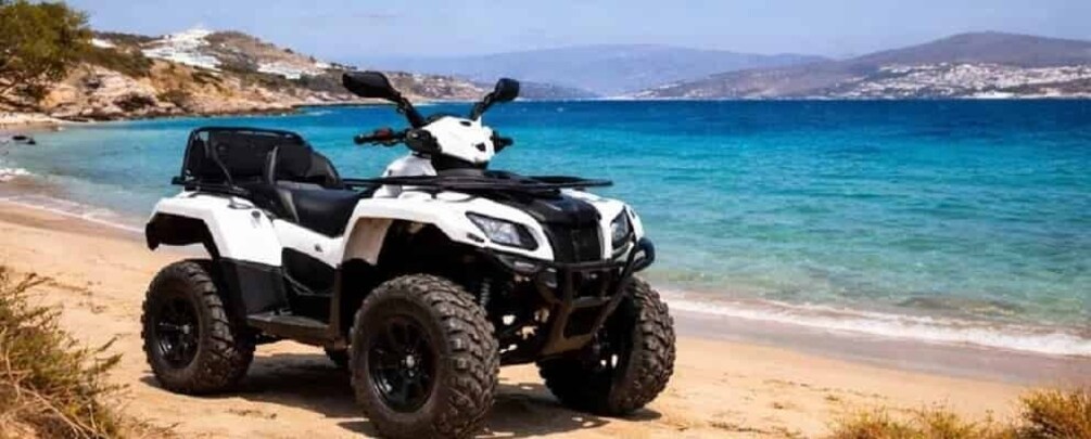 A white ATV on a sandy beach with clear blue water and distant hills. 1