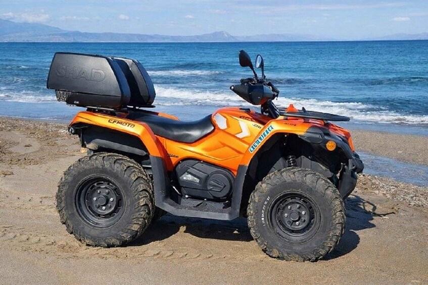 An orange off-road vehicle with a spare tire and a box on the back, parked on a sandy beach with the ocean in the background. 5