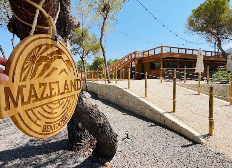 A wooden sign with 'MAZELAND BENIDORM' carved on it, a wooden bridge, and a building with a stone wall in the background. 1