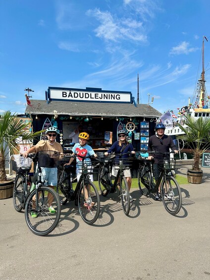 Four people on bicycles in front of a building labeled 'BADUDLEJNING' with a ship in the background. 1
