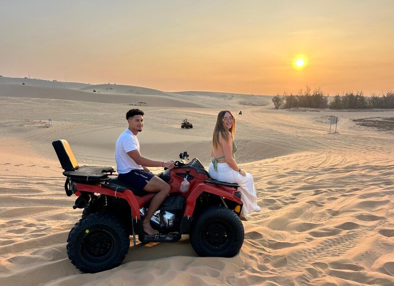 Two people on an all-terrain vehicle in a desert with sand dunes and a sunset. 1