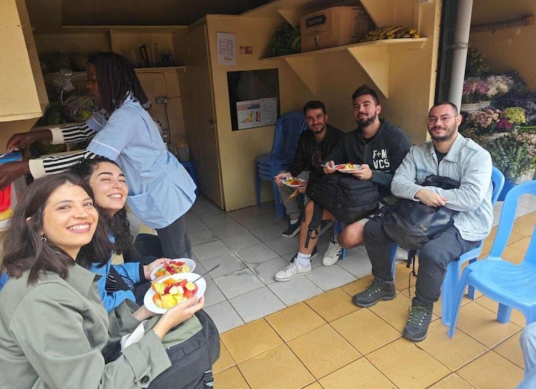 A group of people sitting on plastic chairs, eating plates of food in an indoor setting with tiled floors and a counter in the background. 4