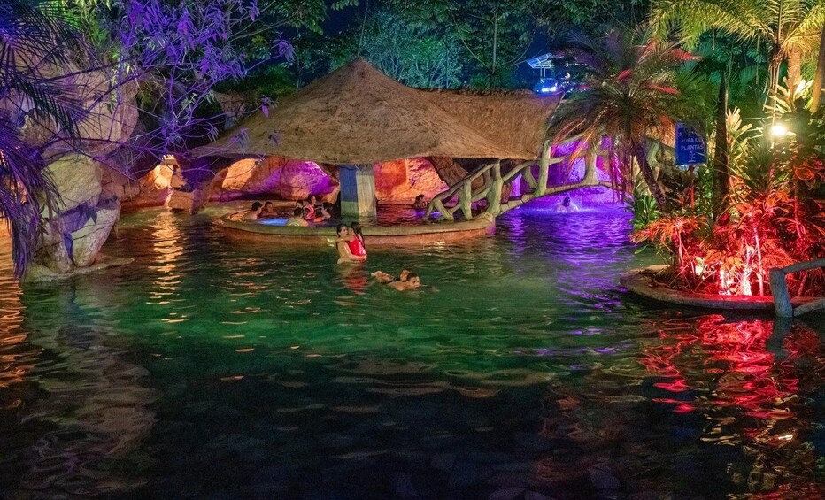 A tropical pool with people swimming, a thatched-roof structure, and colorful lighting. 5