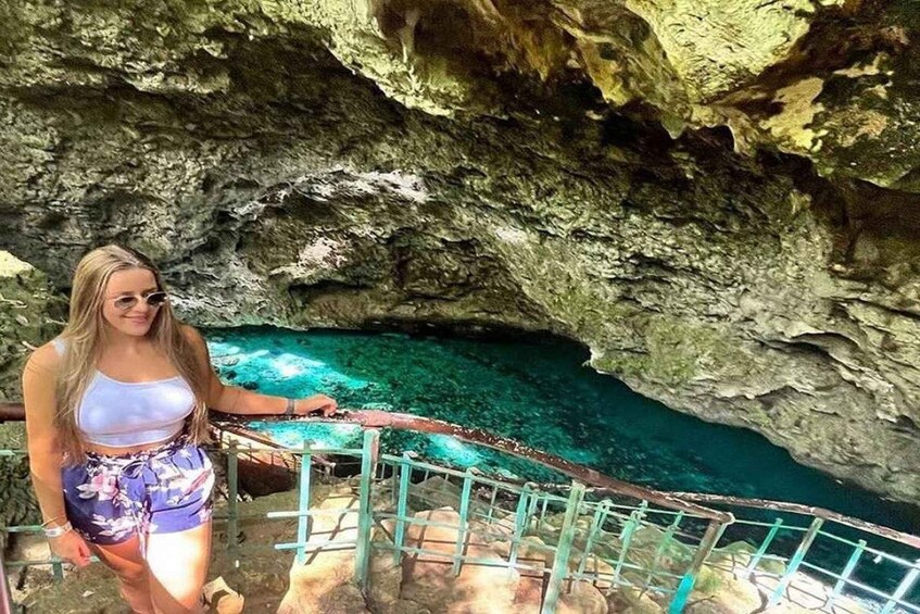 A person standing on a rocky ledge with a railing, overlooking a clear blue pool within a cave. 1