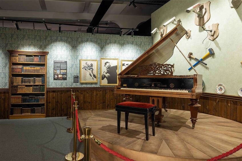 A grand piano, a wooden bookshelf, and framed pictures on a wall in a room with patterned wallpaper. 3