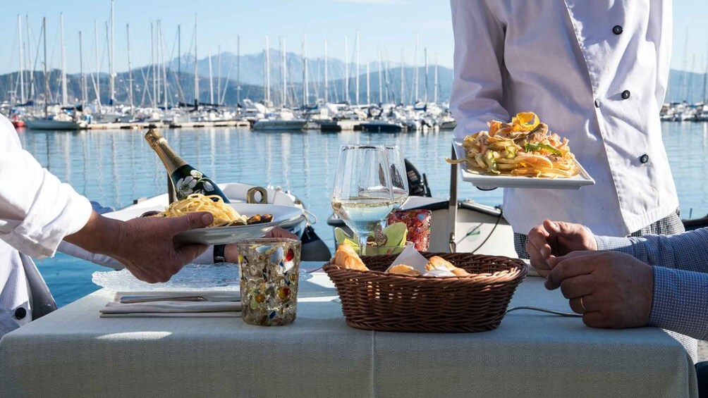 A dining table with food, a bottle of wine, and a glass of white wine at a marina with boats in the background. 2