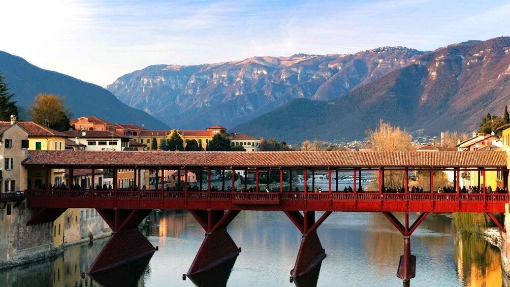 A red bridge over a calm river with buildings and mountains in the background. 1