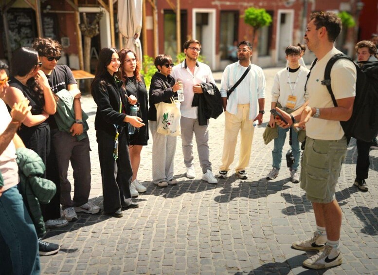 A group of people standing on a cobblestone street, some holding water bottles, in an urban setting with buildings in the background. 2