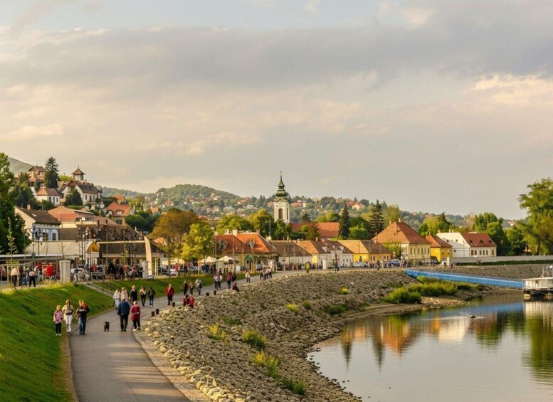 A riverside promenade with people walking, a church with a green spire, and houses with red roofs. 1