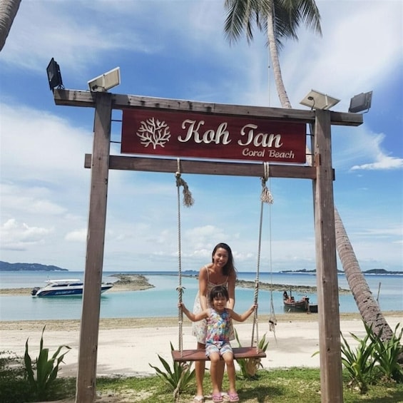 A wooden swing set with a sign reading 'Koh Fan Coral Beach' in front of a beach with palm trees and boats. 5