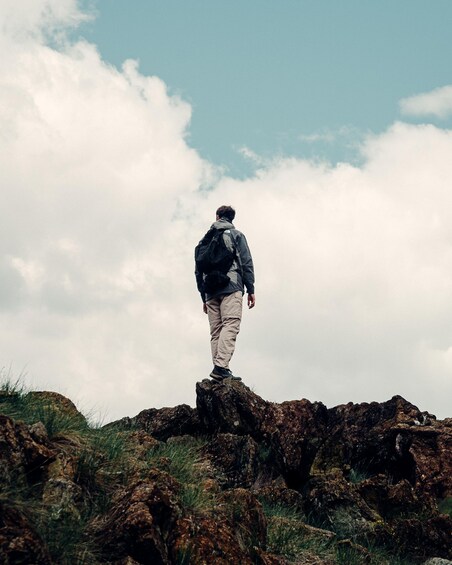 A person standing on a rocky outcrop with a backpack, overlooking a landscape with sparse vegetation and a partly cloudy sky. 1