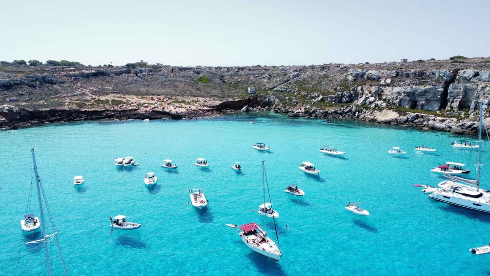 A marina with several boats anchored in clear turquoise water, surrounded by rocky cliffs and a distant town. 2