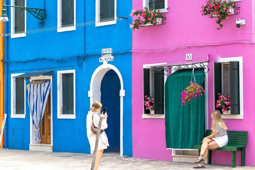 A street with colorful buildings, a person taking a photo, and another person sitting on a bench. 1