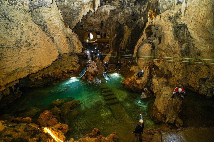 A cave with a clear water pool, stalactites, and people exploring. 4