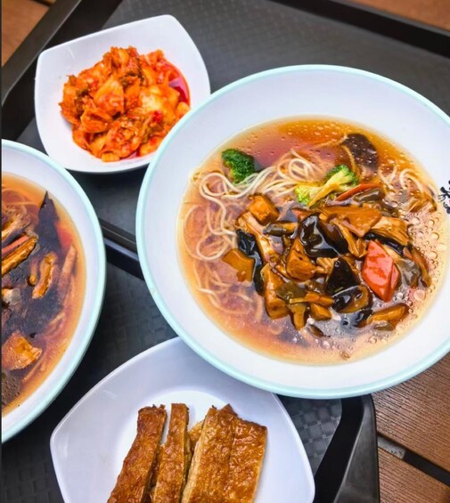A table with three bowls of Asian cuisine, including a noodle soup with vegetables and tofu, a bowl of kimchi, and a plate of fried tofu. 2