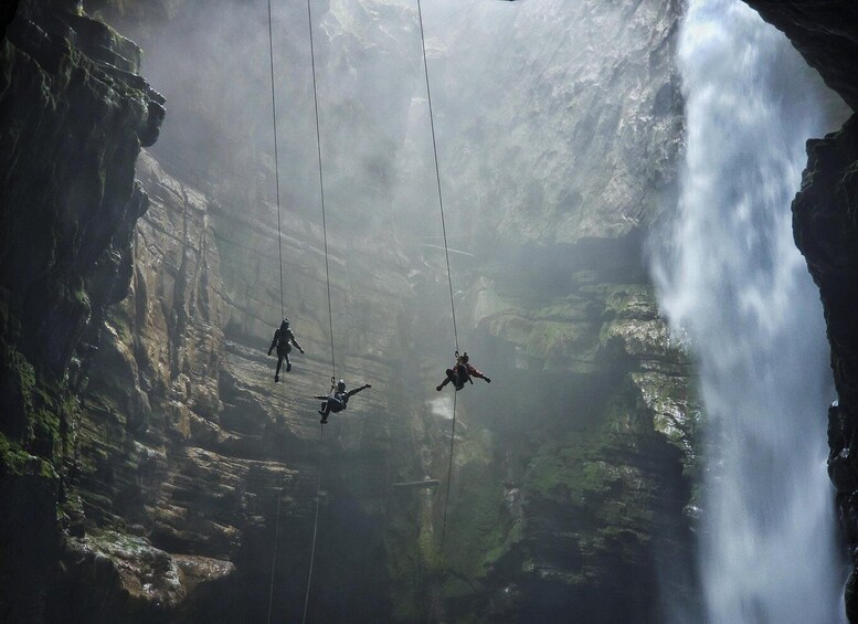 A group of people suspended in mid-air by ropes in a rocky canyon with a waterfall. 1