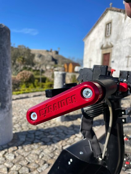 A red handlebar with 'EZRAIDER' engraved on it, a black motorcycle, and a stone-paved area with buildings in the background. 1