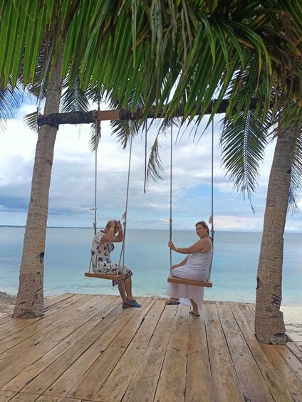 Two people on a wooden swing set with a beach and ocean in the background. 2