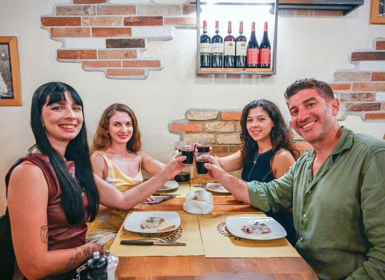 Four people toasting with drinks at a table with plates of food, a wine rack, and a brick wall. 1
