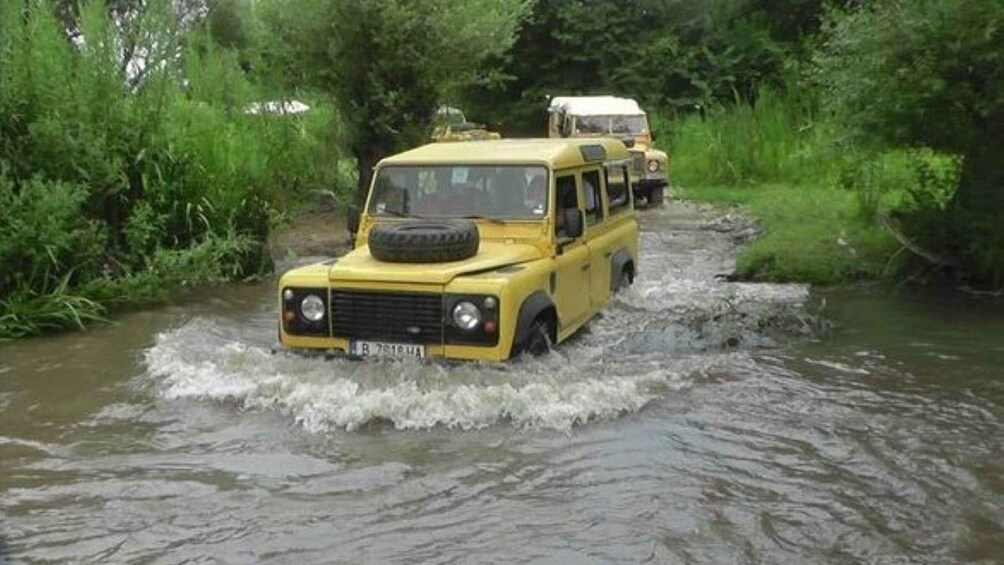 A yellow off-road vehicle crossing a shallow river surrounded by greenery. 2