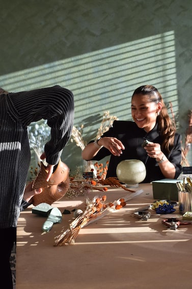 A person holding a small object, a table with various items, and a window with blinds. 2