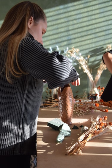 A person cutting a brown object with a knife on a table with dried flowers and a green object. 1