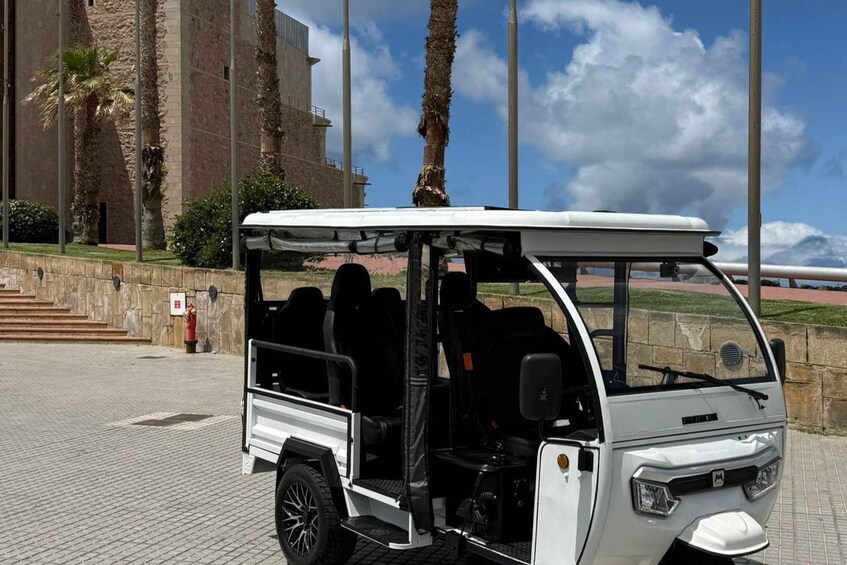 A white off-road vehicle parked on a paved area with palm trees and a stone building in the background. 1