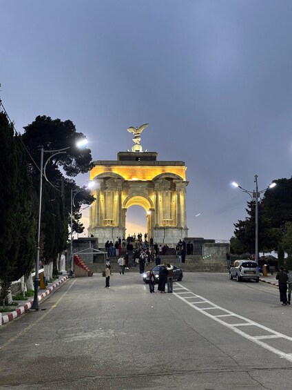 A large illuminated archway with an eagle statue on top, surrounded by people and cars. 5