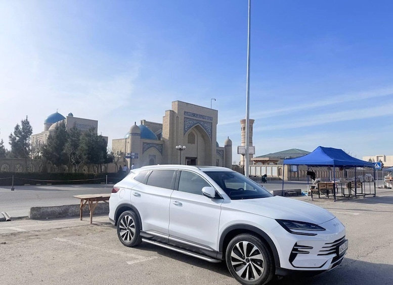 A white SUV parked on a street with a mosque and blue tents in the background. 1