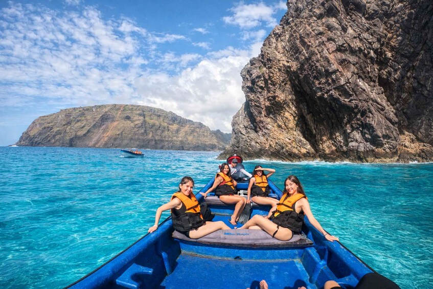 A group of people on a blue boat with life jackets, surrounded by clear turquoise water and a rocky cliff. 1
