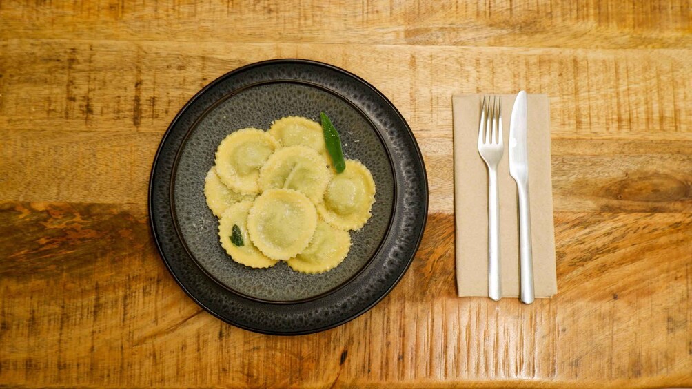 A plate of ravioli with a green garnish on a dark plate, accompanied by a fork and knife on a wooden table. 1