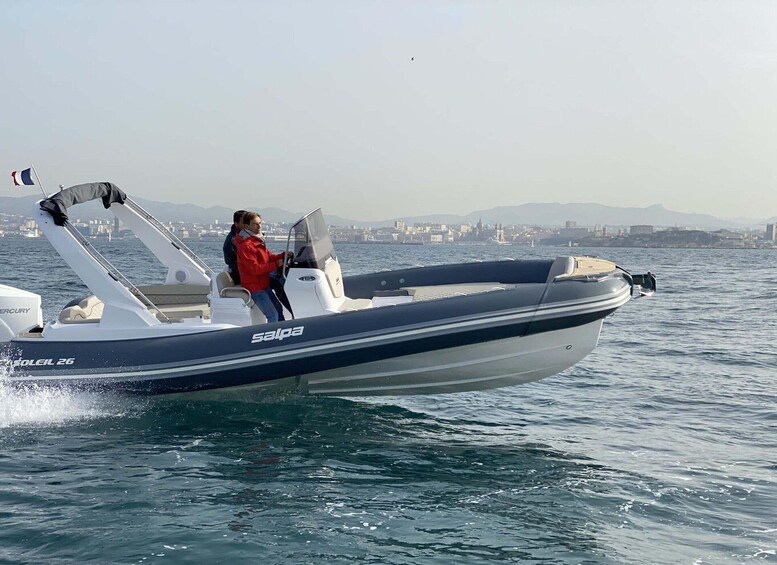 A motorboat with two people on board, moving on a body of water with a cityscape in the background. 1