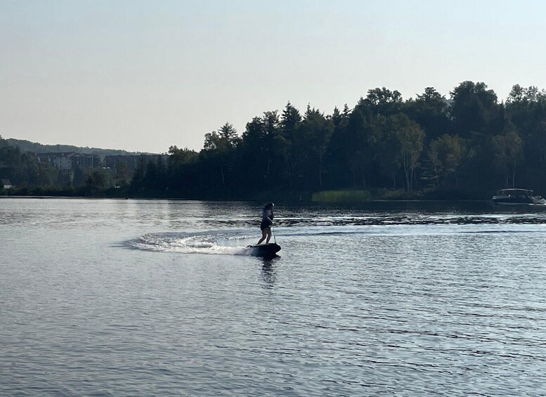 A person wakeboarding on a calm lake with a forested shoreline and a boat in the background. 3