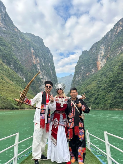 Three people in traditional attire posing by a river with mountainous scenery in the background. 1