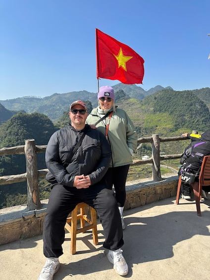 Two people sitting on a bench with a red flag, overlooking a mountainous landscape. 4
