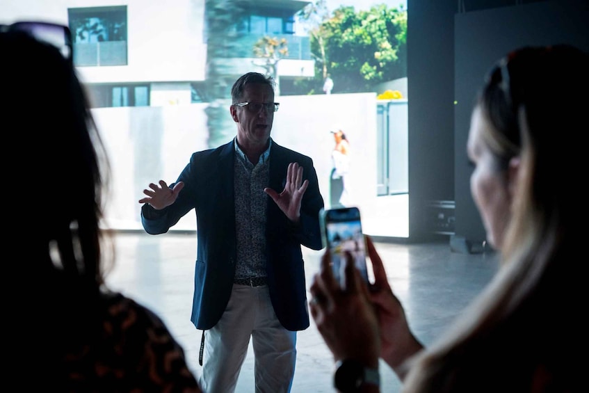 A man in a suit gesturing while speaking to a group of people, with a modern building in the background. 1