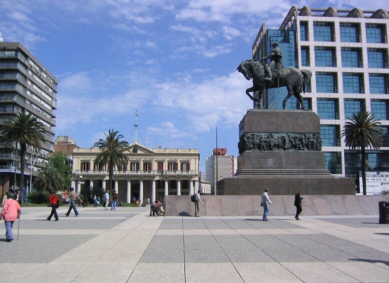 A public square with a statue of a man on horseback, palm trees, and modern buildings. 1