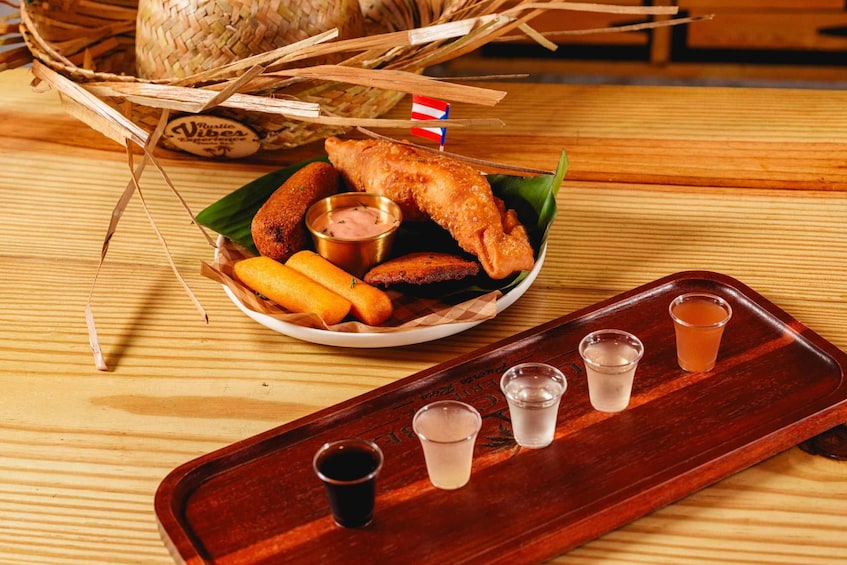A wooden tray with a plate of fried food, including fish and plantains, and four glasses of a clear liquid, possibly a beverage. 1