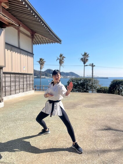 A person in a white martial arts uniform with a black belt, performing a stance outdoors with a traditional building and palm trees in the background. 1