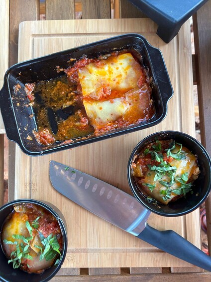 A dish of baked pasta with tomato sauce and melted cheese, garnished with basil, on a wooden board. 2