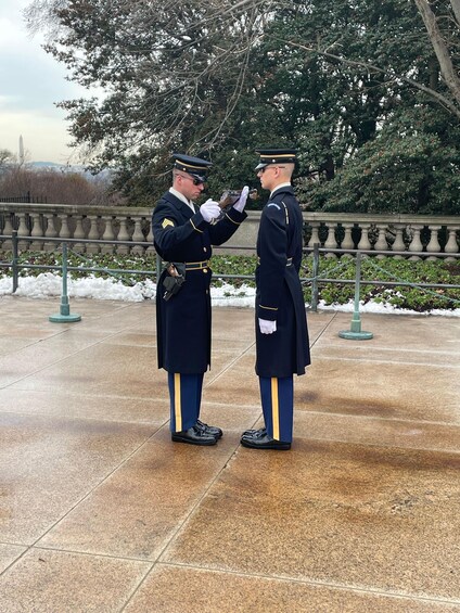 Two uniformed individuals in ceremonial attire standing on a paved area with a balustrade and trees in the background. 1