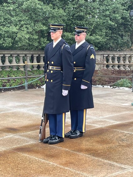 Two uniformed individuals standing in a formal setting with a stone balustrade and greenery in the background. 3